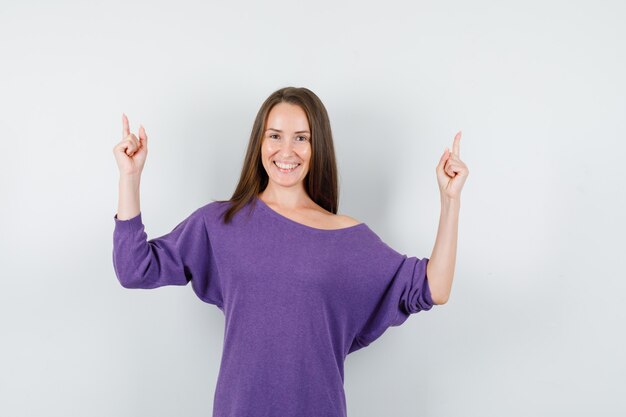 Jeune femme en chemise violette pointant les doigts vers le haut et à la vue de face, heureux.