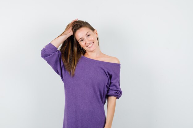 Jeune femme en chemise violette tenant la main dans les cheveux et à la joyeuse vue de face.