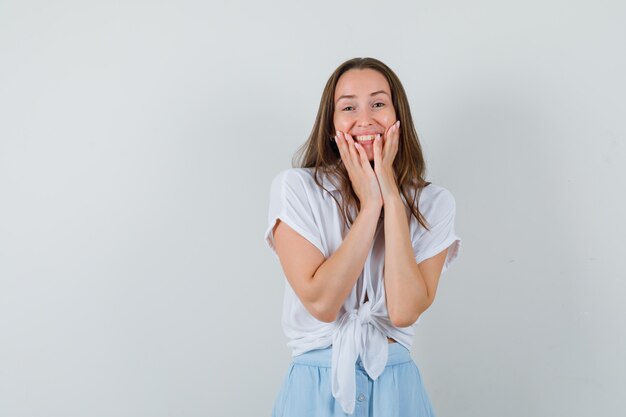 Jeune femme en chemisier blanc et jupe bleu clair tenant les mains sur les joues et à la recherche de plaisir