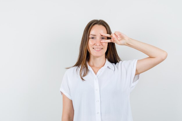 Jeune femme en chemisier blanc montrant le geste gagnant sur ses yeux et à la bonne