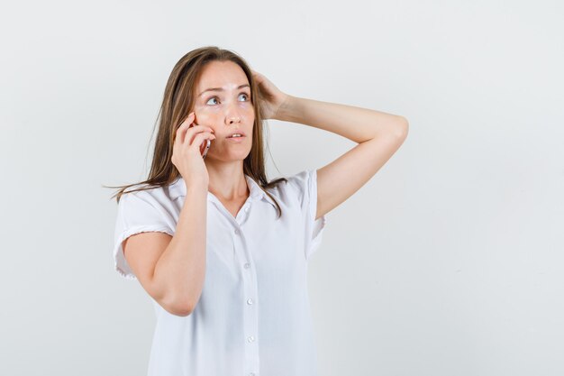 Jeune femme en chemisier blanc parlant au téléphone tout en tenant la main sur sa tête et à la réflexion