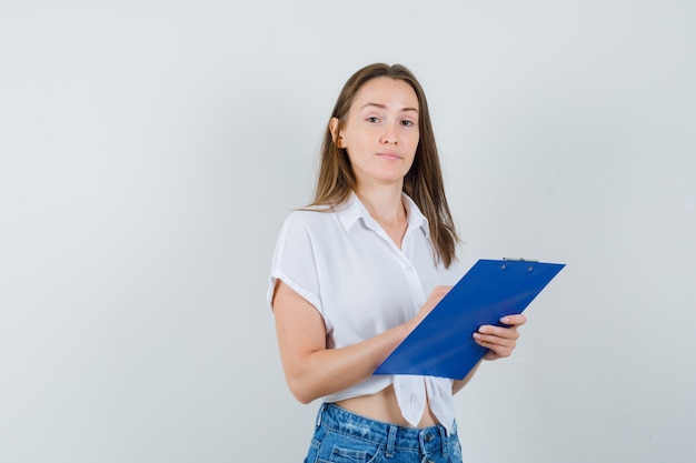 Jeune femme en chemisier blanc tenant le presse-papiers et à la vue attentive, de face.