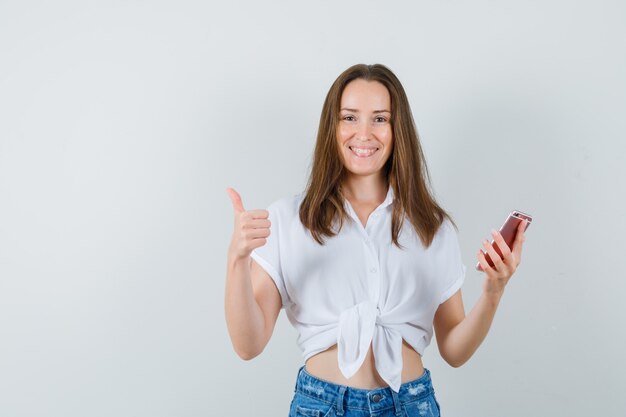 Jeune femme en chemisier blanc tenant le téléphone tout en montrant le pouce vers le haut et à la jolly, vue de face.