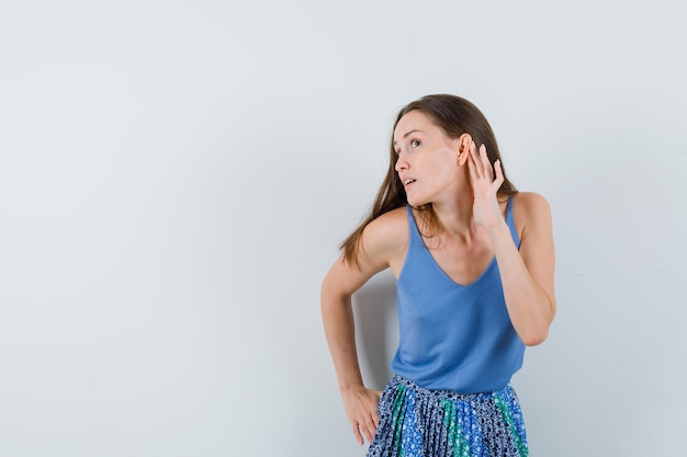 Jeune femme en chemisier bleu, jupe essayant d'écouter quelque chose et regardant attentif, vue de face.