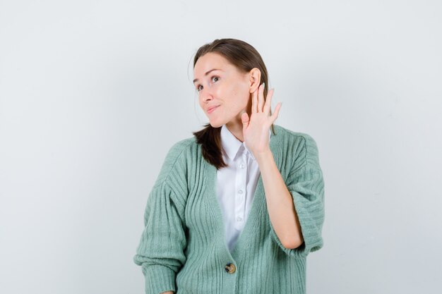 Jeune femme en chemisier, cardigan gardant la main derrière l'oreille et l'air curieux, vue de face.