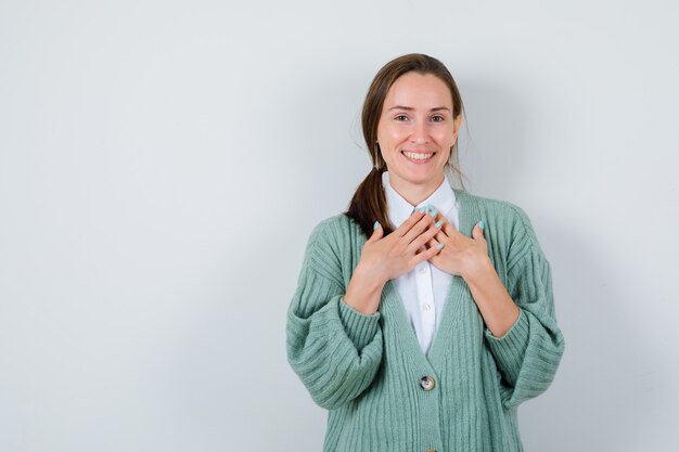 Jeune femme en chemisier, cardigan gardant les mains sur la poitrine et semblant reconnaissante, vue de face.