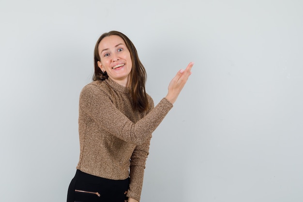 Jeune femme en chemisier doré levant la main tout en montrant quelque chose sur le côté gauche et à la vue de face, heureux. espace pour le texte