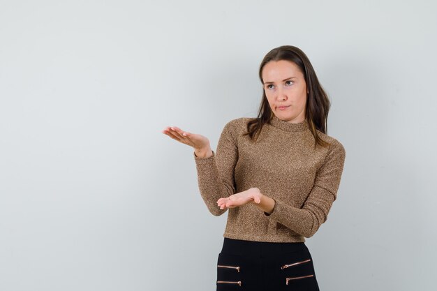 Jeune femme en chemisier doré, levant les mains avec une manière interrogative et à la recherche pensive, vue de face.