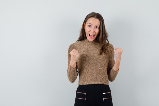 Jeune femme en chemisier doré montrant le geste du gagnant et à la vue de face, énergique. espace pour le texte