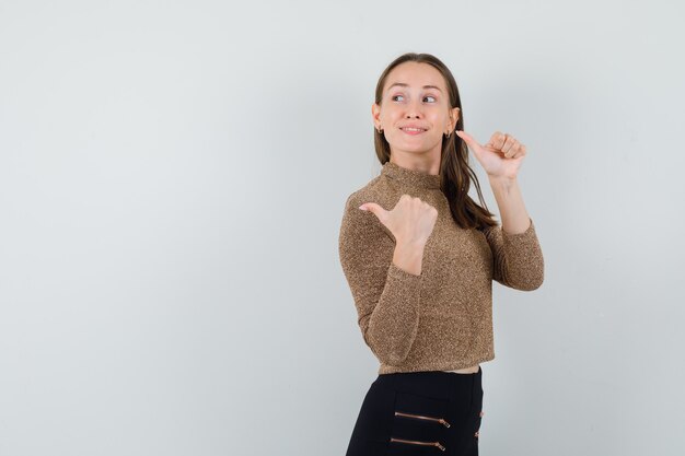 Jeune femme en chemisier doré regardant en arrière tout en montrant le pouce vers le haut et à la joie. espace pour le texte