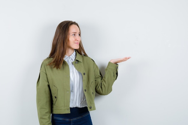 Jeune femme en chemisier écartant la paume de la main et semblant jolie, vue de face.