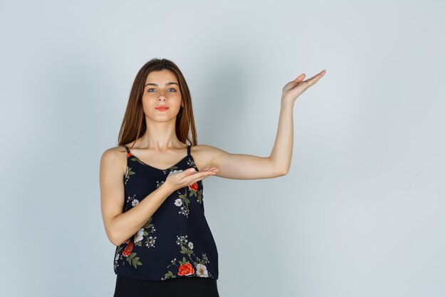 Jeune femme en chemisier, jupe montrant un geste de bienvenue et l'air confiant, vue de face.