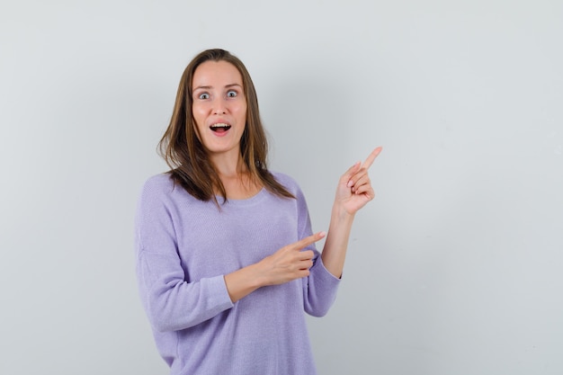 Jeune femme en chemisier lilas pointant vers le coin supérieur gauche tout en ouvrant largement les yeux et à la surprise