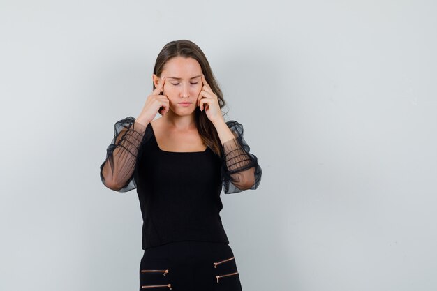 Jeune femme en chemisier noir et pantalon noir frottant les tempes et à la vue épuisée, de face.