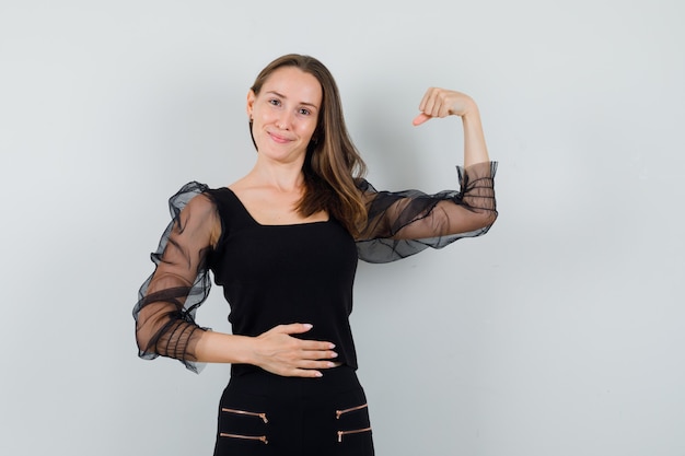 Photo gratuite jeune femme en chemisier noir et pantalon noir montrant ses muscles et tenant une main sur le ventre et à l'optimiste