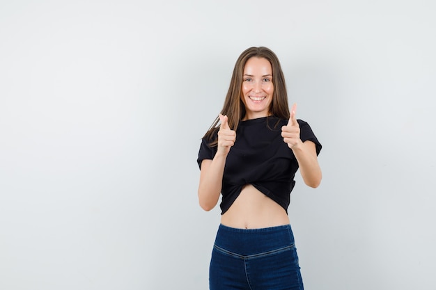 Jeune femme en chemisier noir, pantalon pointant vers la caméra avec le geste du pistolet et à l'optimiste