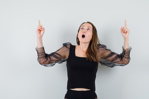 Jeune femme en chemisier noir regardant avec la mâchoire tombée tout en pointant vers le haut et à la surprise