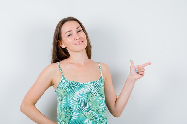 Jeune femme en chemisier pointant vers le côté droit et semblant joyeuse, vue de face.