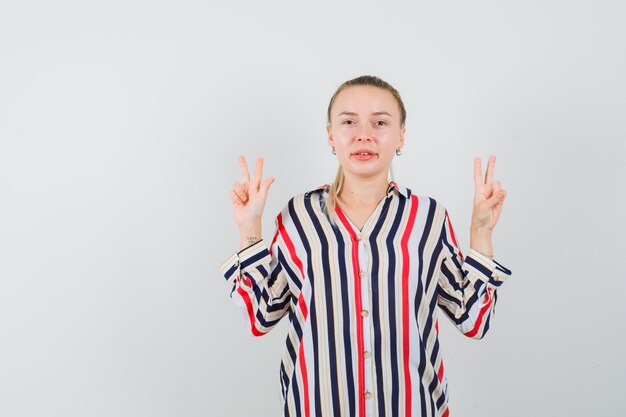 Jeune femme en chemisier rayé montrant le geste de paix avec ses deux mains et à l'optimiste