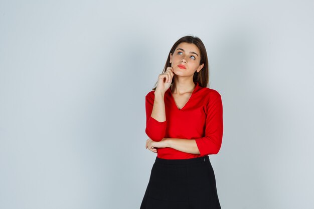 Jeune femme en chemisier rouge, jupe debout dans la pose de réflexion et à la pensif