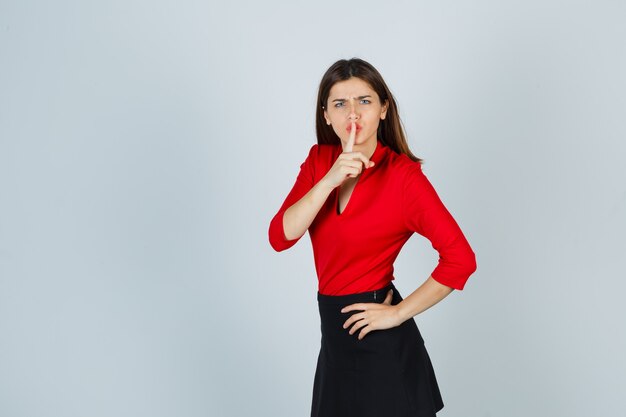 Jeune femme en chemisier rouge, jupe noire montrant le geste de silence