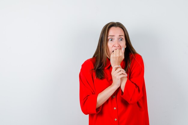 Jeune femme en chemisier rouge avec le poing sur la bouche et l'air effrayé, vue de face.