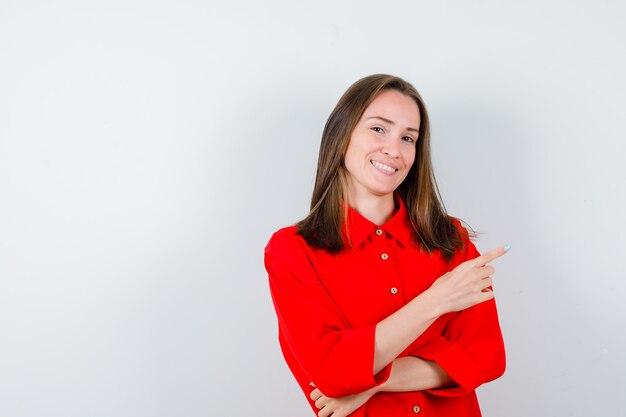 Jeune femme en chemisier rouge pointant vers le haut et l'air heureux, vue de face.