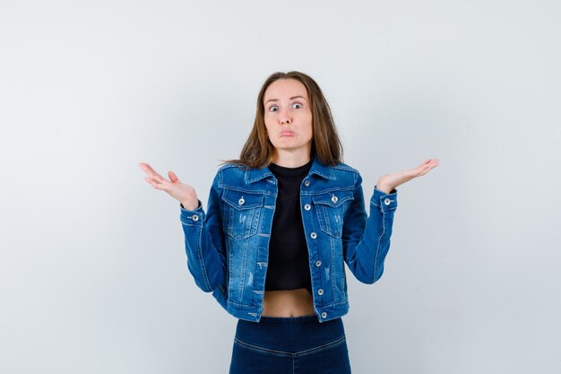 Jeune femme en chemisier, veste, jeans montrant un geste impuissant et l'air confus, vue de face.