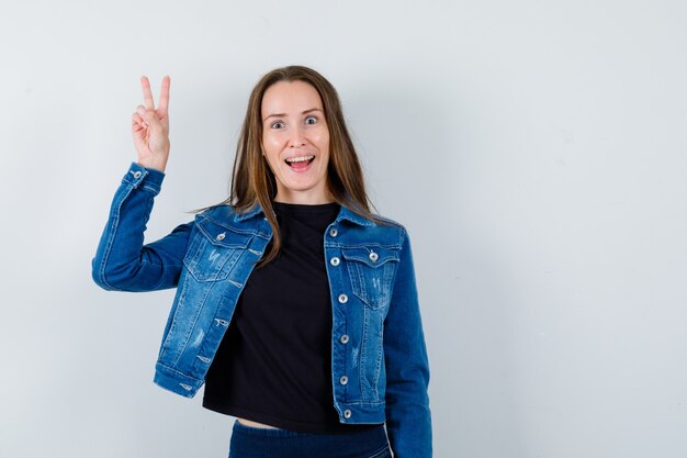 Jeune femme en chemisier, veste montrant le signe V et semblant joyeuse, vue de face.