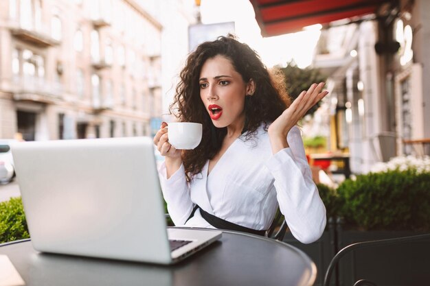 Jeune femme choquée aux cheveux bouclés noirs assise à la table avec une tasse de café à la main et regardant étonnamment sur un ordinateur portable dans un café dans la rue