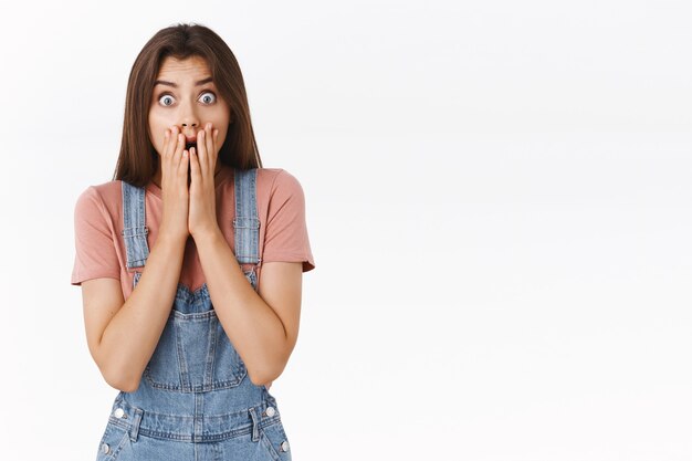 Une jeune femme choquée, inquiète et inquiète, apprenant des nouvelles dévastatrices, la bouche au toucher haletant et les yeux éclatants impressionnés, debout sans voix sur fond blanc, réagissant nerveusement