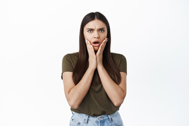 Jeune femme choquée et inquiète préoccupée par les mauvaises nouvelles, fronçant les sourcils et haletant, se tenant la main sur les joues, s'inquiétant pour la personne, debout contre un mur blanc