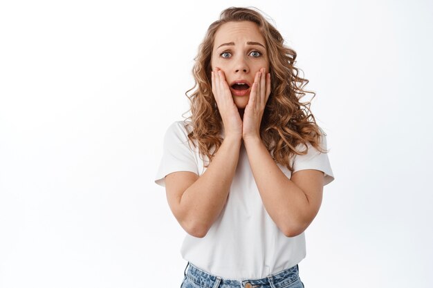 Jeune femme choquée en panique, se tenant la main sur le visage, criant et ayant l'air inquiète, exprime de la pitié et des émotions nerveuses, debout sur un mur blanc