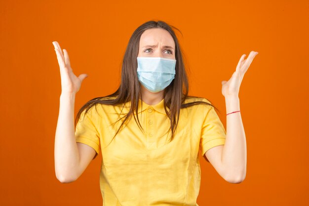 Jeune femme choquée en polo jaune et masque de protection médicale haussant les épaules de panique sur fond orange isolé