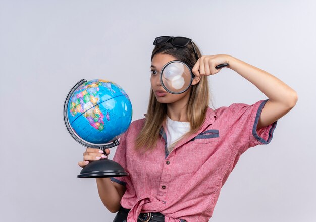 Une jeune femme choquée portant une chemise rouge à lunettes de soleil tenant un globe tout en le regardant avec une loupe sur un mur blanc