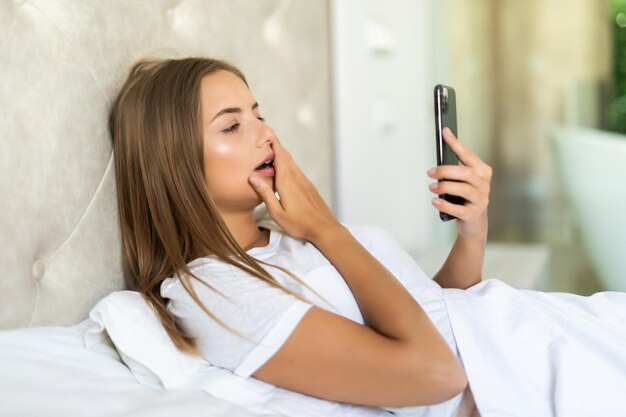 Jeune femme choquée regardant un téléphone portable dans son lit à la maison