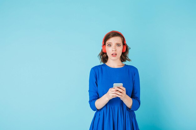 Jeune femme choquée en robe et casque rouge debout avec un téléphone portable et regardant étonnamment à huis clos sur fond bleu