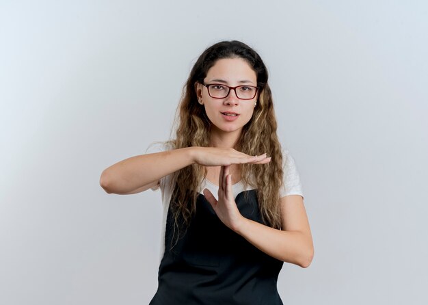 Jeune femme de coiffeur professionnel en tablier à l'avant faisant le geste de temps avec les mains debout sur un mur blanc