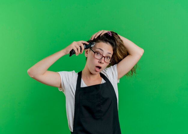 Jeune femme de coiffeur professionnel en tablier de couper ses cheveux avec tondeuse à l'avant d'être surpris debout sur le mur vert