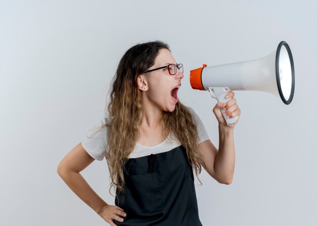 Jeune femme de coiffeur professionnel en tablier criant au mégaphone bruyamment debout sur un mur blanc