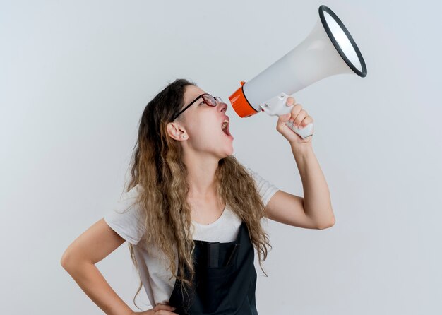 Jeune femme de coiffeur professionnel en tablier criant au mégaphone bruyamment debout sur un mur blanc