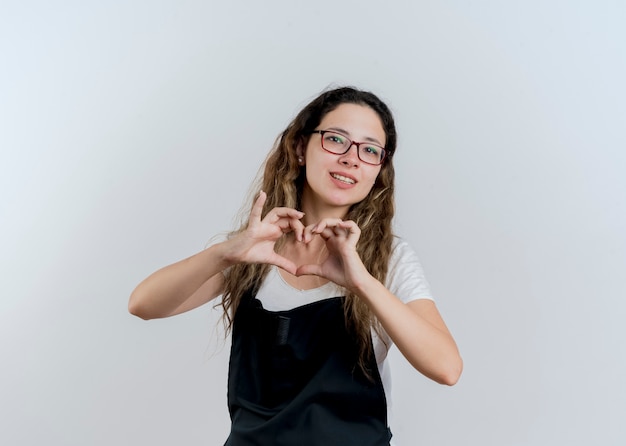 Jeune femme de coiffeur professionnel en tablier faisant le geste du cœur avec les doigts souriant debout sur un mur blanc