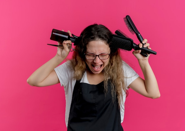 Jeune femme de coiffeur professionnel en tablier holding spray et brosses à cheveux à la frustration de cris avec une expression agressive