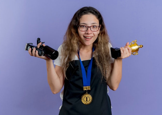 Photo gratuite jeune femme de coiffeur professionnel en tablier avec médaille d'or autour du cou tenant tondeuse et trophée souriant avec visage heureux