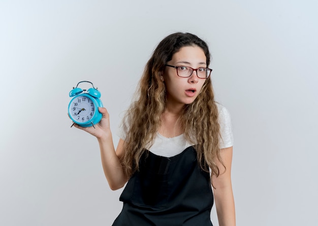 Photo gratuite jeune femme de coiffeur professionnel en tablier montrant un réveil à l'avant surpris debout sur un mur blanc