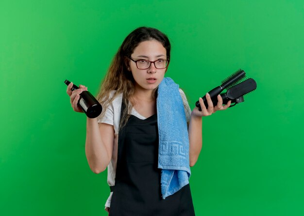 Jeune femme de coiffeur professionnel en tablier avec une serviette sur l'épaule tenant des brosses à cheveux et spray à côté avec visage sérieux