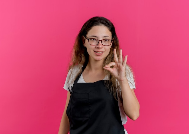 Jeune femme de coiffeur professionnel en tablier souriant faisant signe ok