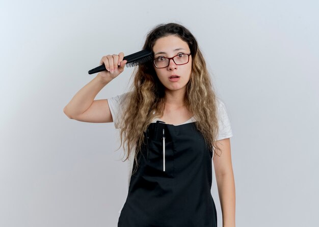 Jeune femme de coiffeur professionnel en tablier tenant une brosse à cheveux peignant ses cheveux à l'avant confus debout sur un mur blanc