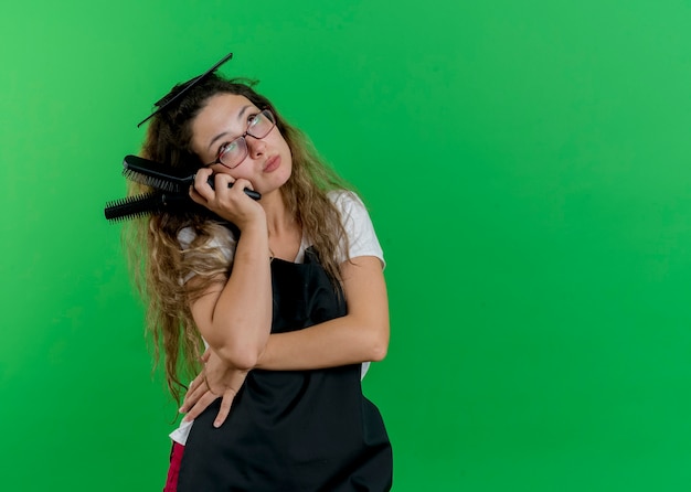 Jeune femme de coiffeur professionnel en tablier tenant des brosses à cheveux à la perplexité