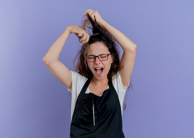 Jeune femme de coiffeur professionnel en tablier tenant des ciseaux essayant de couper ses cheveux en criant avec une expression agacée debout sur un mur bleu